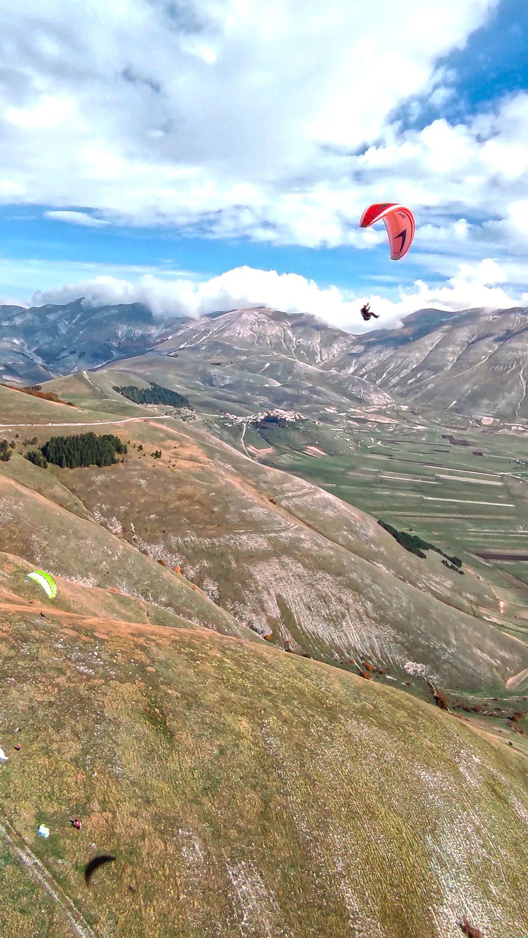 Luftaufnahme: Am Boden sieht man den Startplatz Veletta, wo sich andere Piloten vorbereiten und in der Luft zwei andere Gleitschirme.